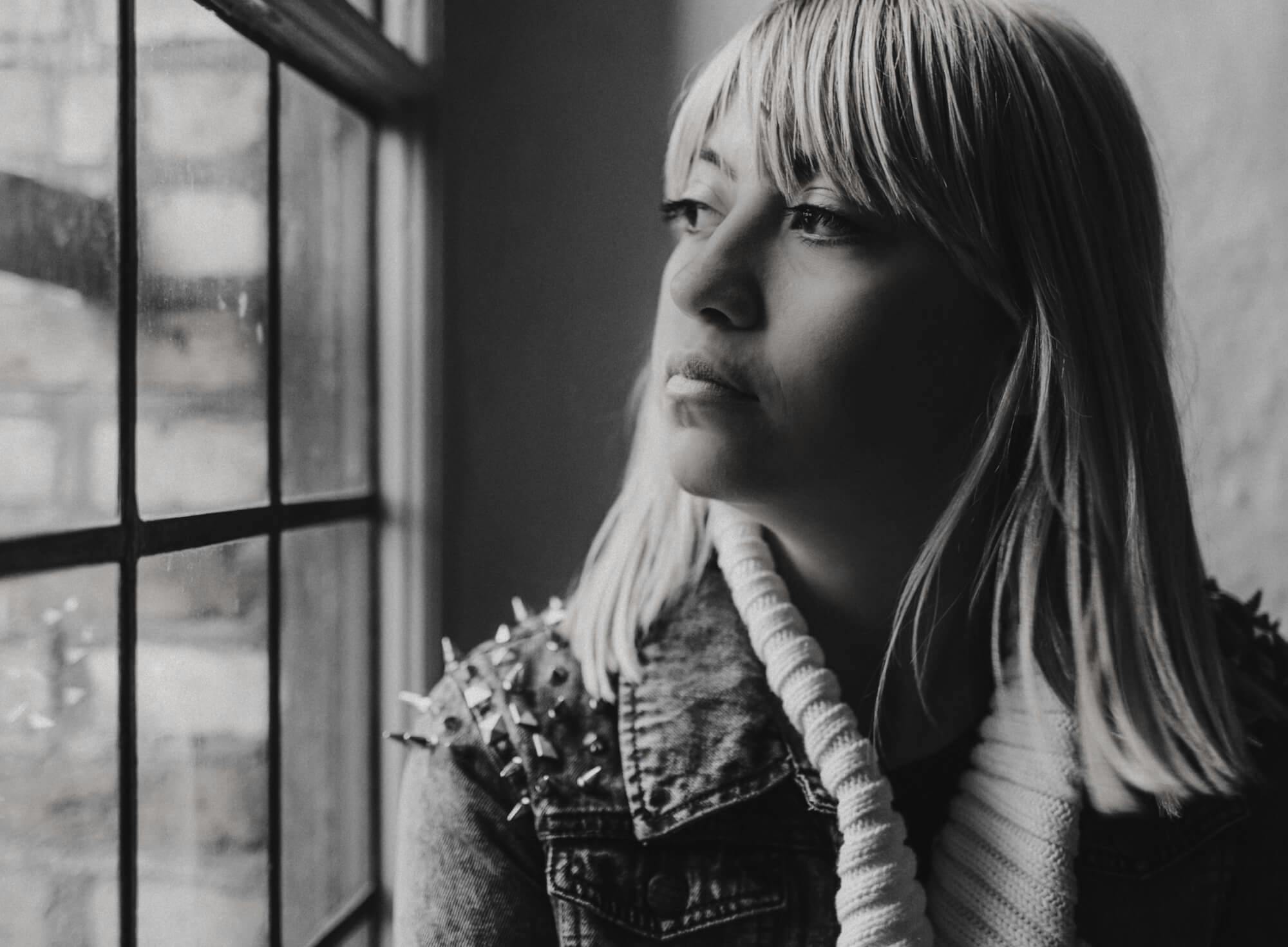 A black & white photo of a woman staring solemnly out a window.