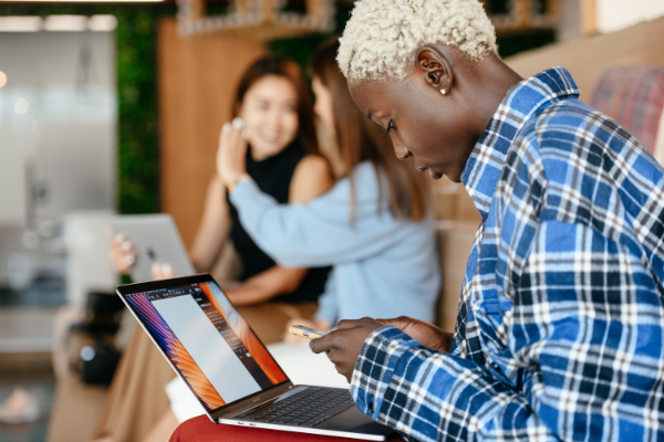 A young woman wearing a plaid shirt looking at a phone and laptop with two young women talking in the background