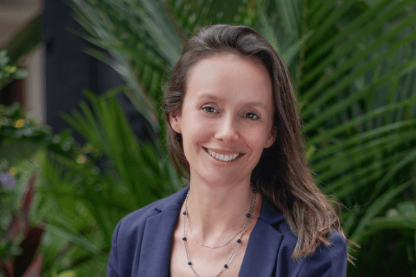 A headshot of a woman with brown hair wearing a blazer, plants in background